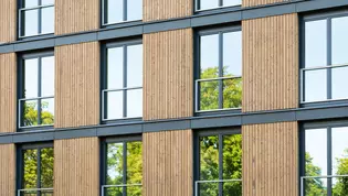 Die Abbildung zeigt eine Holzfassade an einem Bürogebäude mit Baumspiegelung in den Fenstern.